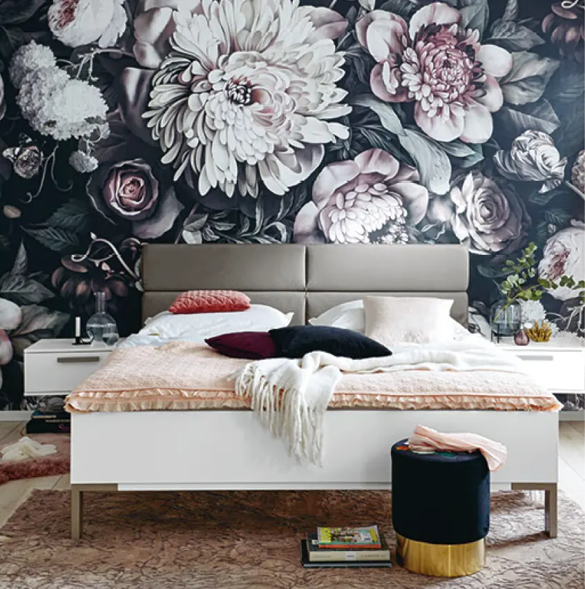Weisses Bett vor dunkler Blumenwand, gemusterter Teppich, zwei Nachttische, Kissen in Rosa und Schwarz, runder Hocker mit goldfarbenem Sockel